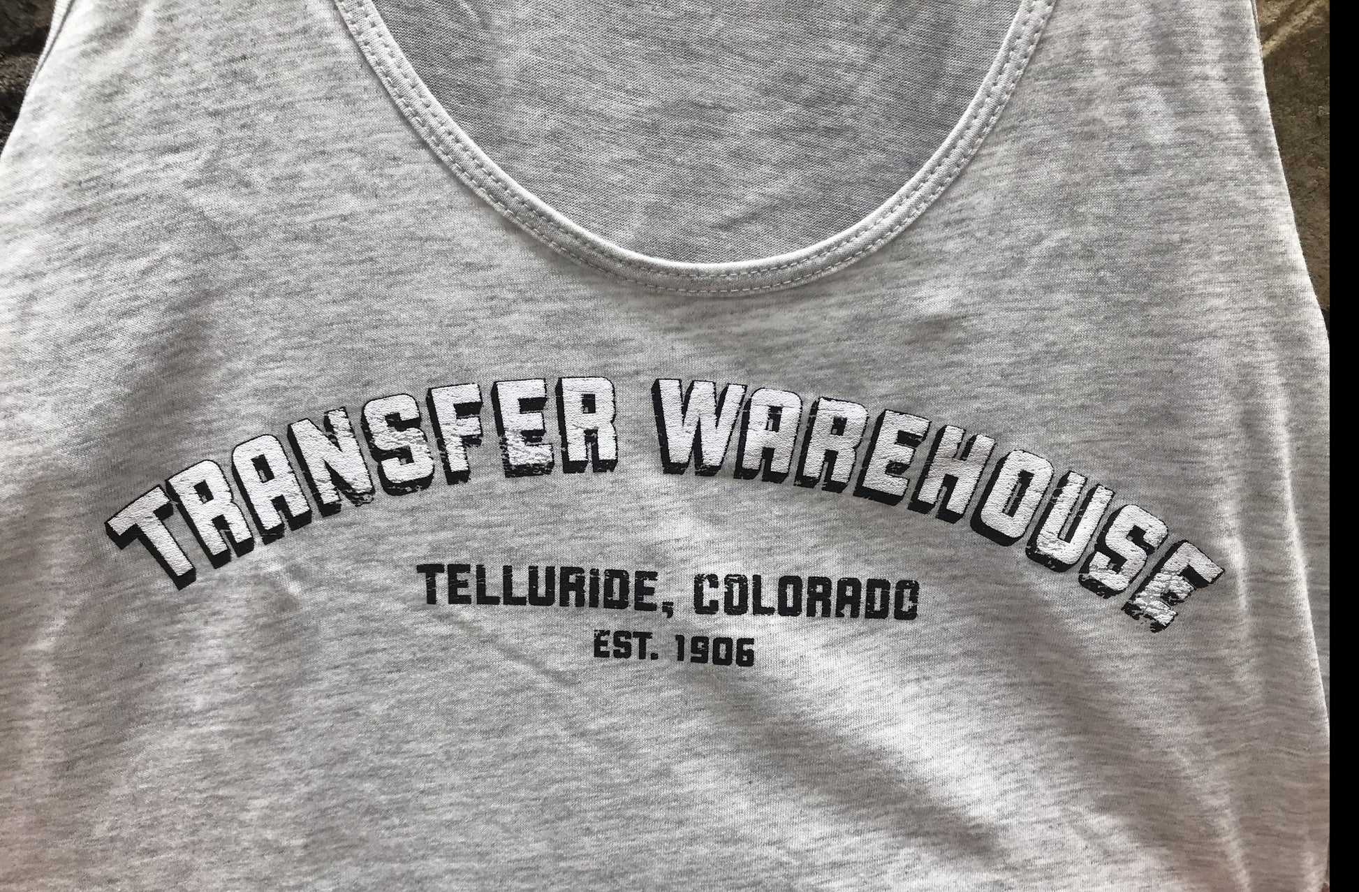 Light Grey tank top with the words "Transfer Warehouse. Telluride, Colorado. Est. 1906." Close up on words and fabric details.